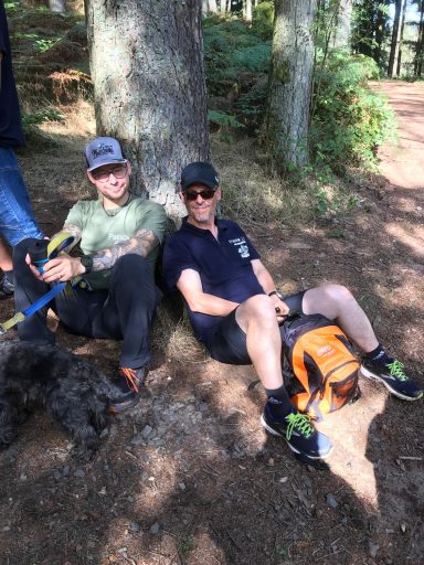 Zwei Männer sitzen an einen Baum gelehnt, in einem Waldgebiet, entspannen sich.