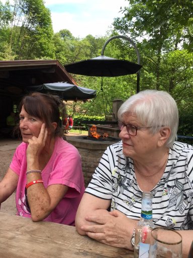 Zwei Frauen sitzen an einem Tisch im Freien, umgeben von Bäumen und Natur.