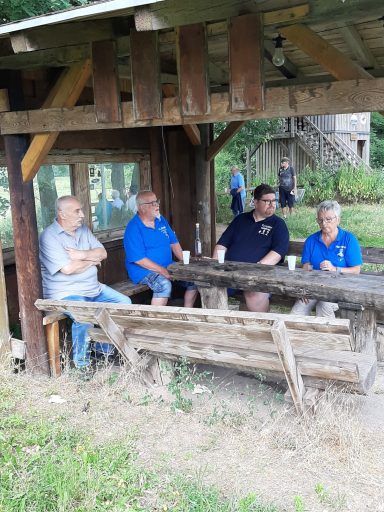 Vier Männer sitzen an einem Tisch unter einer Holzpergola, umgeben von Natur.