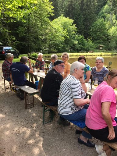 Gruppe von Menschen sitzt an einem See, im Hintergrund Bäume und ein ruhiger Weg.