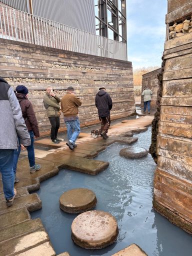 Gruppe von Personen geht über Steine in einem Wassergang zwischen Mauern.