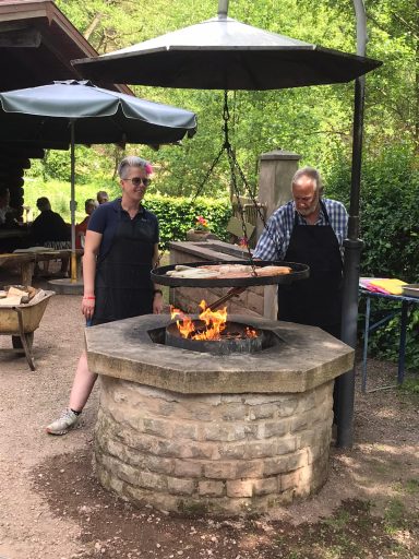Zwei Personen stehen an einem Feuerplatz, umgeben von Bäumen und Sitzplätzen.