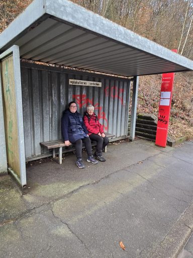 Zwei Personen sitzen an einer Bushaltestelle in einer ländlichen Umgebung.