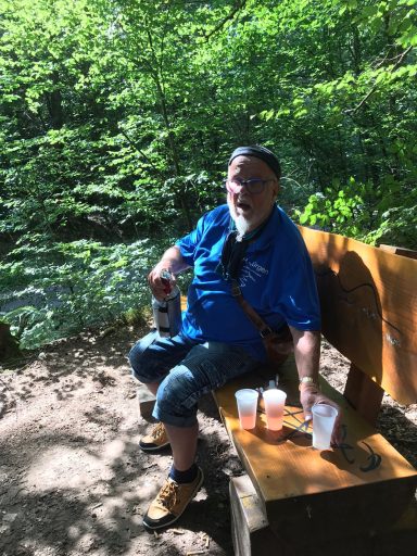 Mann in blauer Kleidung sitzt entspannt auf einer Bank im Wald mit Getränken neben sich.