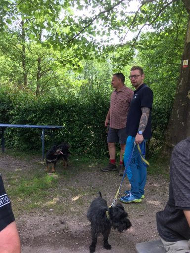 Zwei Männer mit Hunden stehen im Grünen an einem Tisch.