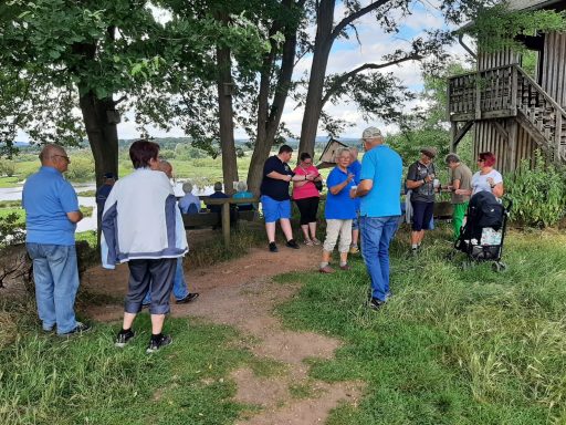 Gruppe von Menschen steht unter Bäumen, während einige miteinander sprechen und essen.