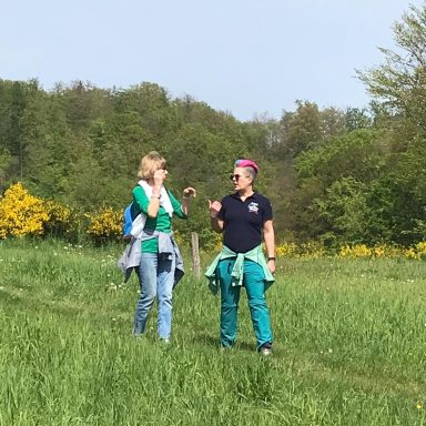 Zwei Frauen stehen auf einem grünen Feld und unterhalten sich, umgeben von Bäumen und Sträuchern.