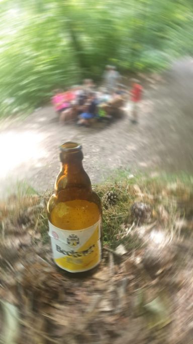 Braune Flasche auf dem Boden, verschwommene Personen im Hintergrund im Wald.