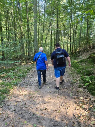 Zwei Personen gehen einen Waldweg entlang, umgeben von Bäumen und grüner Vegetation.