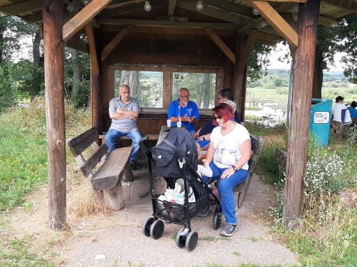 Vier Personen sitzen an einer Holzbank unter einem Überdach in einer landschaftlichen Umgebung.