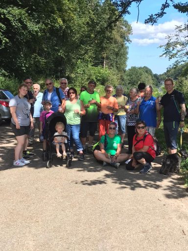 Gruppe von Menschen im Freien, einige mit Kindern, umgeben von Bäumen und Autos.