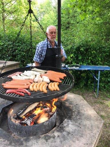 Ein Mann grillt verschiedene Würstchen über offenem Feuer in einem Garten.