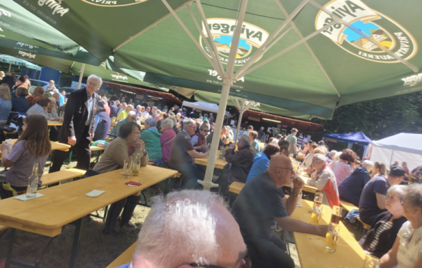 Menschenmenge in einem Biergarten mit langen Bänken und Tischen unter einem Sonnendach.