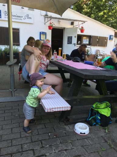 Kleines Kind steht an einem Tisch, umgeben von mehreren Personen im Freien.