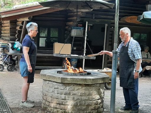 Zwei Personen stehen an einem Feuerplatz und bereiten Essen zu.