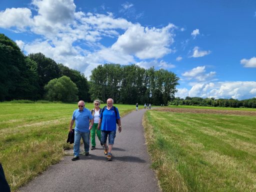 Zwei Männer wandern auf einem Weg entlang einer grünen Wiese unter blauem Himmel.