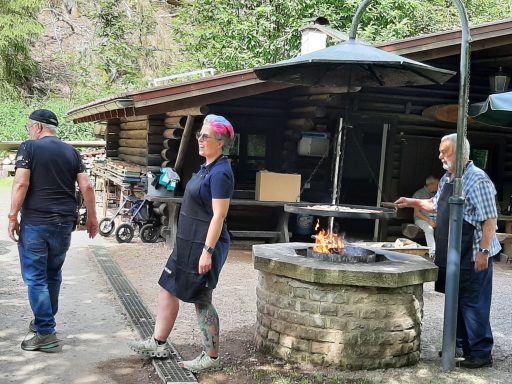 Drei Personen am Grill im Freien, umgeben von Bäumen und einem Holzhaus.