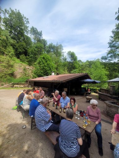Gruppe von Menschen sitzt an einem Tisch im Freien, umgeben von Natur und Bäumen.