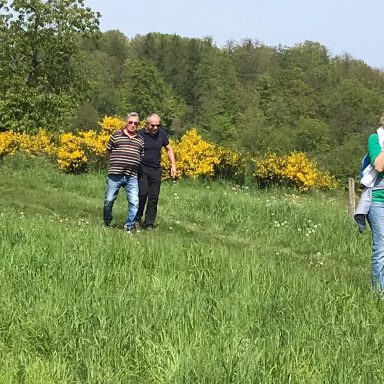 Zwei Männer gehen auf einer Wiese, umgeben von gelben Blumen und grüner Landschaft.