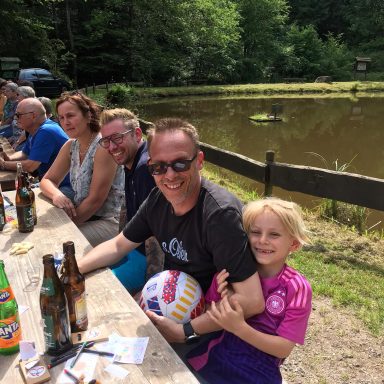 Eine fröhliche Gruppe von Menschen sitzt an einem Tisch am Wasser, Kinder lachen und spielen.