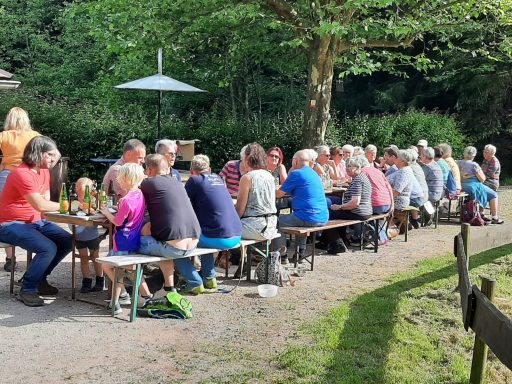 Gruppen von Menschen sitzen an langen Bänken unter Bäumen und genießen eine Mahlzeit im Freien.