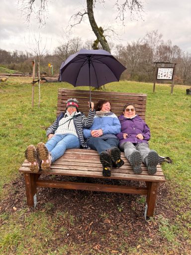 Drei Personen liegen entspannt auf einer Holzliege unter einem Regenschirm im Freien.