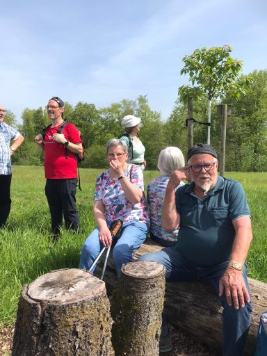 Gruppe von Menschen im Freien, einige sitzen auf Baumstämmen, andere stehen.