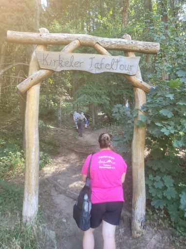 Eingang zu einem Wanderweg, Holzschild mit Aufschrift, umgeben von Bäumen und Wanderern.
